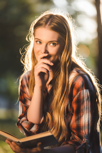 portrait shot of beautiful girl in casual clothes sitting on meadow in sunlight, holding book and looking away - Foto, Imagem