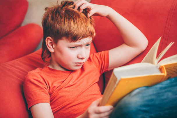 The red-haired boy is very puzzled by what he read in the book and looks at the pages with perplexity - Foto, imagen