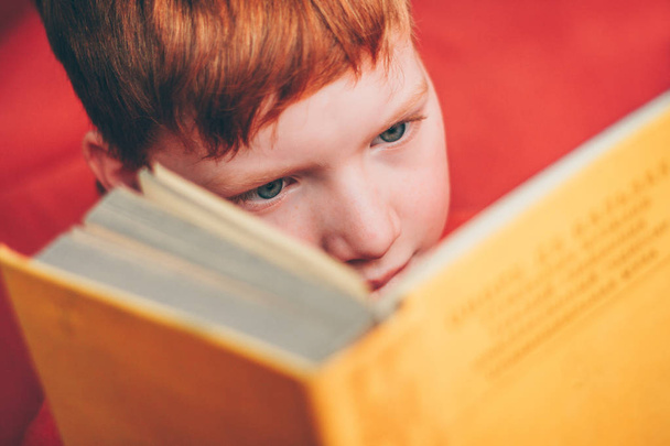 Redheaded child carefully reads adventure novel, close-up of emotion on face - Фото, изображение