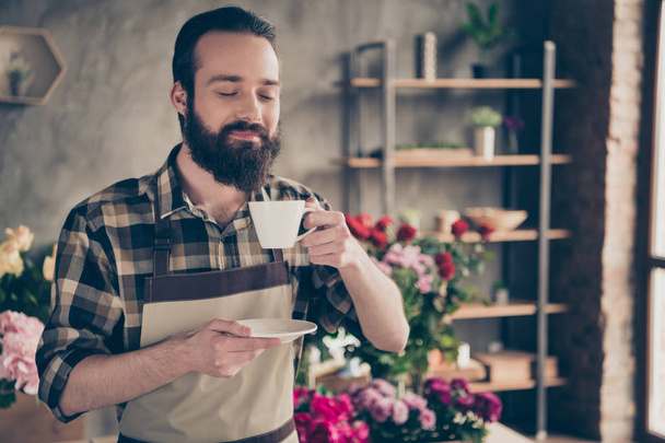 Close-up portrait of his he nice attractive cheerful guy wearing uniform drinking hot morning espresso among peony bunch retail at industrial concrete style salon indoors - Foto, immagini