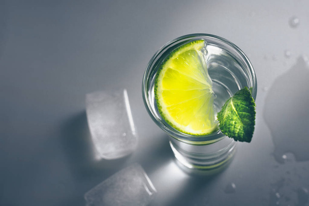 Top view of a shot of vodka with a lime slice and ice on a glass table - Фото, изображение