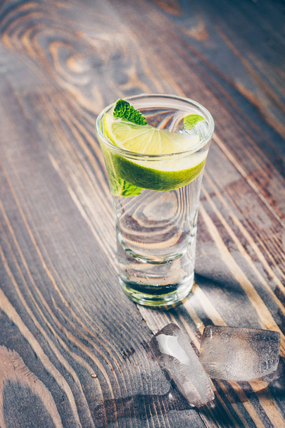 Cold tequila in a glass on a wooden table with a slice of lime, mint and ice slices - Fotó, kép