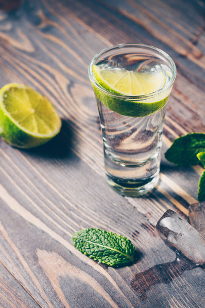 Cold vodka in a shot glass on a wooden table with a slice of lime, mint and ice slices - Фото, изображение