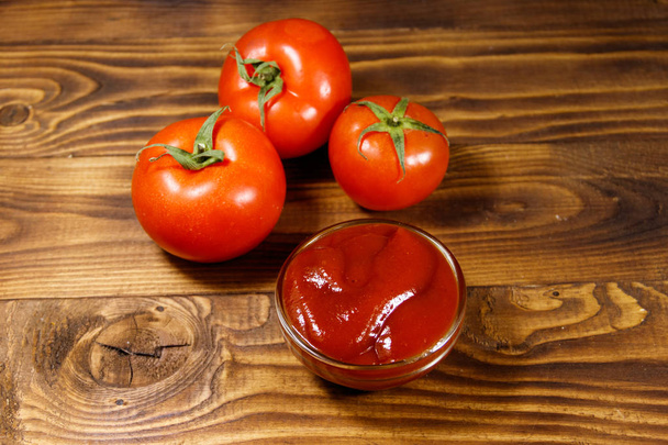 Glass bowl of ketchup or tomato sauce and fresh ripe tomatoes on wooden table - Photo, Image