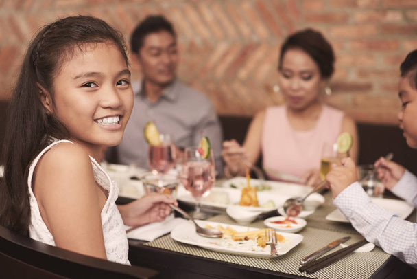 Портрет милой улыбающейся девушки, сидящей за столом со своей семьей за ужином в ресторане
 - Фото, изображение