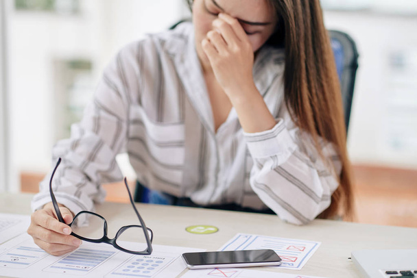 Mujer joven con exceso de trabajo sosteniendo anteojos y frotando los ojos mientras está sentada en el escritorio en la oficina
 - Foto, Imagen