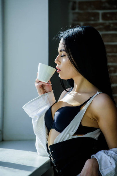 Enjoying morning sunlight. Sensual girl sexy breasts drinking coffee near windowsill. Attractive female lingerie relaxing at home. Passionate mysterious lover. Perfect coffee time. But first coffee - Фото, зображення