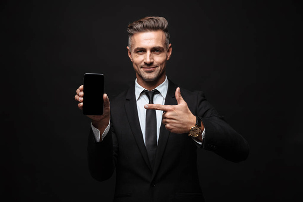 Портрет дорослого впевненого бізнесмена, одягненого в офіційний костюм посміхаючись і вказуючи палець на мобільний телефон
 - Фото, зображення