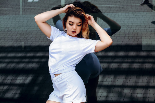 Κομψό κορίτσι με ένα λευκό μπλουζάκι και λευκό σορτς στέκεται κοντά στο κτίριο. - Φωτογραφία, εικόνα
