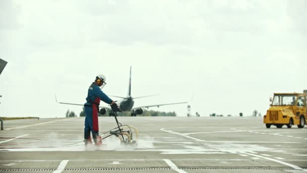 A man worker slowly cleaning the asphalt on the airport field using water machine - a plane passing by - Footage, Video