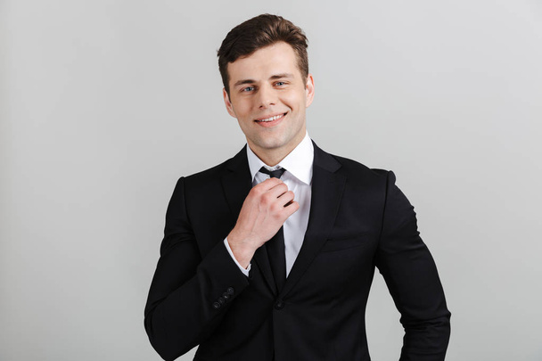 Image of confident young businessman in formal suit touching his tie and smiling at camera - Foto, imagen