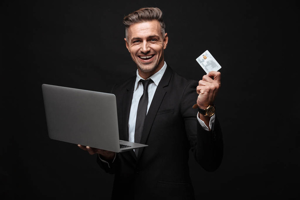 Portrait of manly smiling businessman dressed in formal suit holding laptop computer and credit card - 写真・画像
