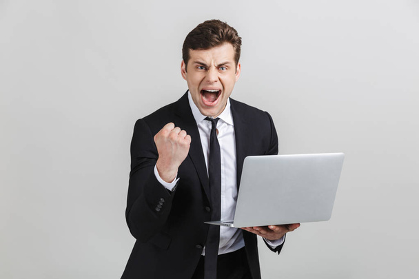 Image of successful excited businessman in formal suit celebrating triumph while holding laptop isolated over gray background - Fotoğraf, Görsel