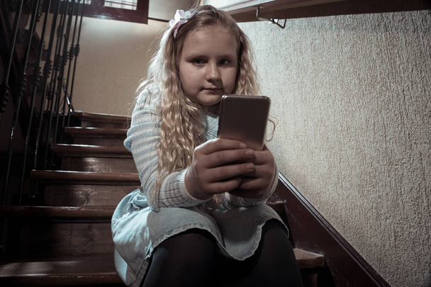 Sad depressed young girl victim of cyberbullying by mobile smart phone sitting on stairs feeling lonely, unhappy, hopeless and abused. Child bullied and harassed by text message by online stalker. - Fotografie, Obrázek