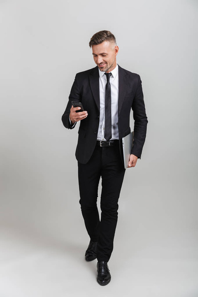 Photo of handsome adult businessman in formal suit smiling while holding laptop and cellphone - Fotografie, Obrázek