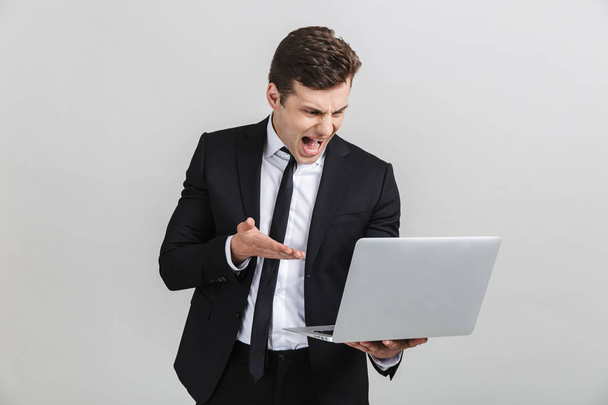Portrait of stressed irritated young businessman in office suit screaming while holding laptop - Foto, imagen