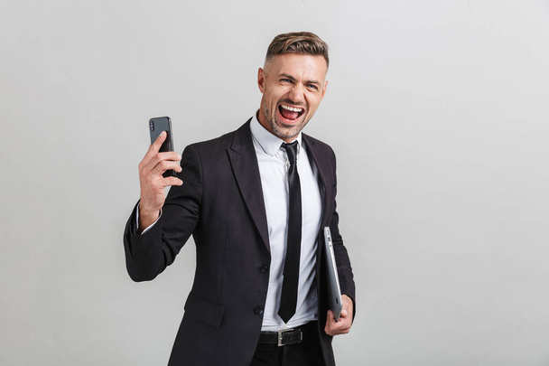 Portrait of ecstatic adult businessman in office suit holding smartphone and laptop while standing - Фото, изображение