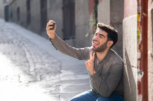 Excited trendy young man taking selfie or blogging on smart mobile phone on holidays in european city street or university campus. Holidays and technology Connection and social media concept. - 写真・画像