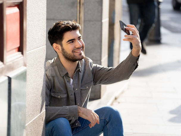 Excited trendy young man taking selfie or blogging on smart mobile phone on holidays in european city street or university campus. Holidays and technology Connection and social media concept. - Photo, Image