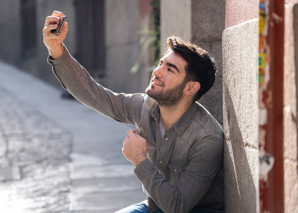 Excited trendy young man taking selfie or blogging on smart mobile phone on holidays in european city street or university campus. Holidays and technology Connection and social media concept. - Foto, Imagen