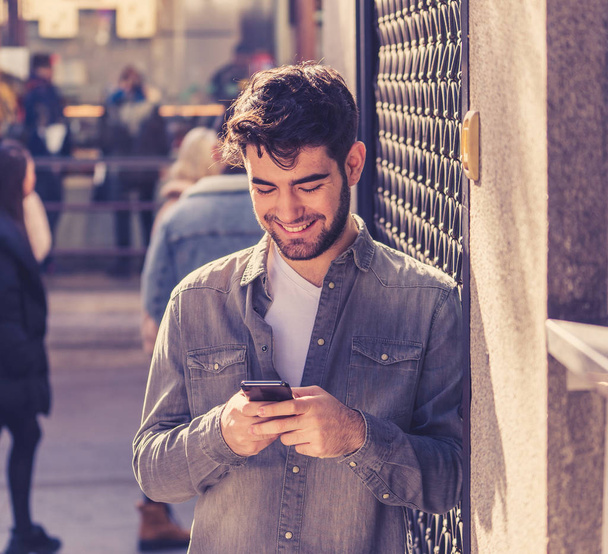 Fashion college man in his twenties happy checking blog or chatting on the internet with friends on smart mobile phone app in european outdoors city. In communication technology and travel Europe. - 写真・画像