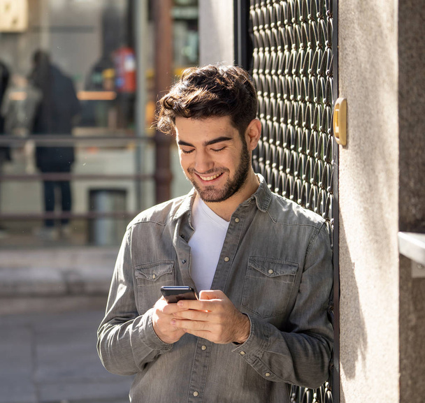 Μόδα κολλέγιο άνδρας στο τριάντα χαρούμενος έλεγχος blog του ή chat στο διαδίκτυο με τους φίλους για την έξυπνη εφαρμογή κινητού τηλεφώνου στην Ευρωπαϊκή ύπαιθρο πόλη. Στην τεχνολογία επικοινωνίας και τα ταξίδια στην Ευρώπη. - Φωτογραφία, εικόνα