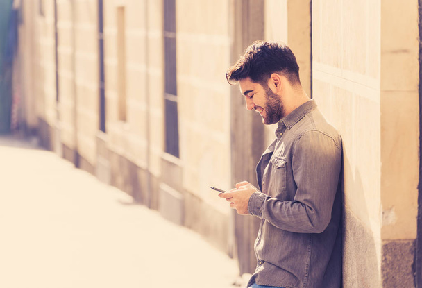Fashion college man in his twenties happy checking blog or chatting on the internet with friends on smart mobile phone app in european outdoors city. In communication technology and travel Europe. - 写真・画像