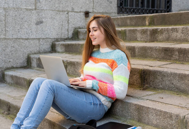 Attractive trendy teen student or young college girl working on laptop on the internet, blogging, chatting and checking email on steps in a european city. In Student learning online City lifestyle. - Фото, зображення