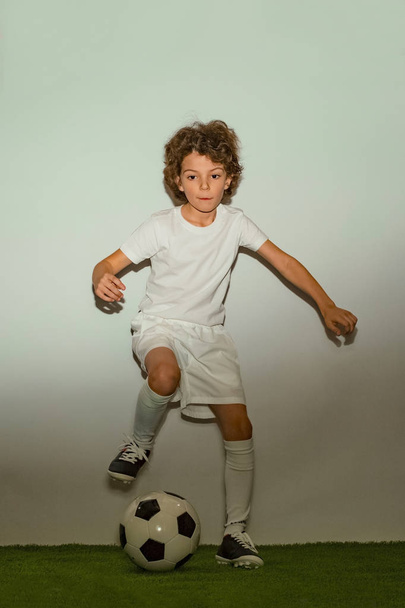 Young boy as a soccer or football player in sportwear making a feint or a kick with the ball for a goal on white studio background. Fit playing boy in action, movement, motion at game. - Photo, Image