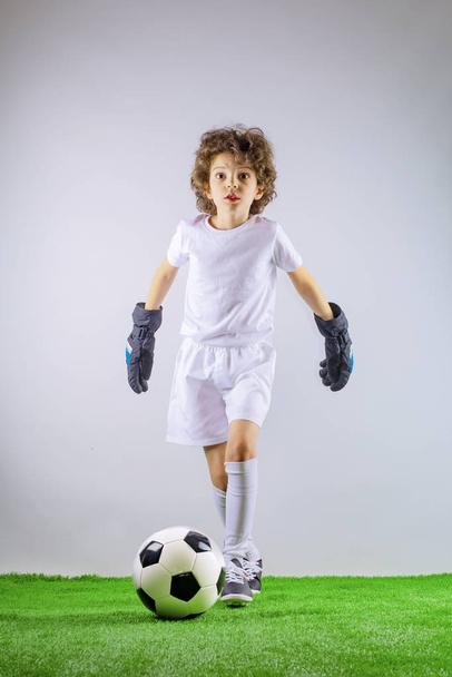 Young boy as a soccer or football player in sportswear standing with the ball like a winner, the best forward or goalkeeper on a gray background. Fit playing boy in action, movement, motion at game. - Φωτογραφία, εικόνα