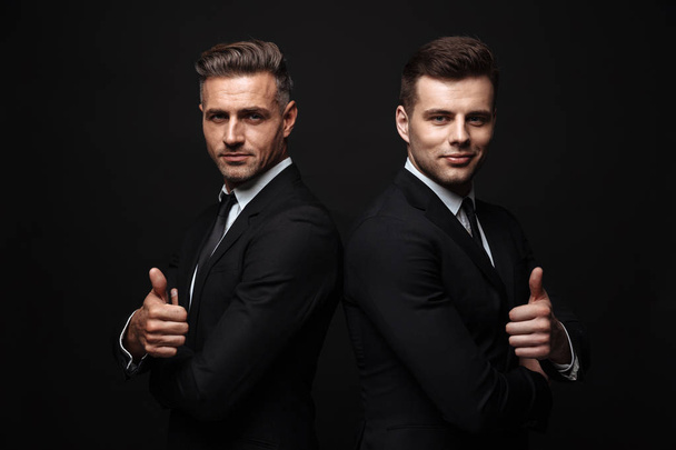 Portrait of smiling two businessmen dressed in formal suit posing at camera back to back with thumb up - Photo, image