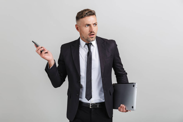 Portrait of outraged adult businessman in office suit holding smartphone and laptop while standing - 写真・画像