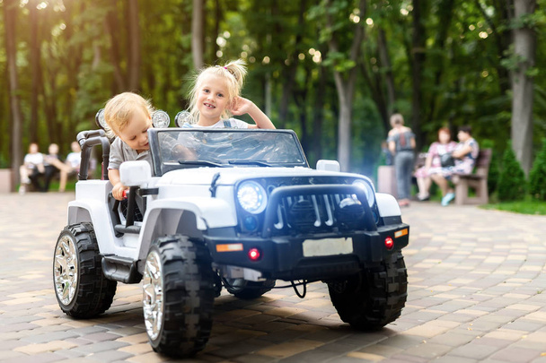 2つのかわいいかわいいブロンドは、市内の公園で電気おもちゃのSUV車に乗って楽しみを持っている子供たちをサイビング。兄弟姉妹は、屋外の街路で遊び、運転車両を楽しんでいます。幸せな子供時代の概念 - 写真・画像