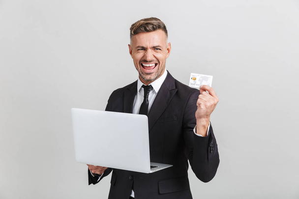 Image of positive adult businessman in formal suit rejoicing while holding laptop and credit card - Foto, Imagen