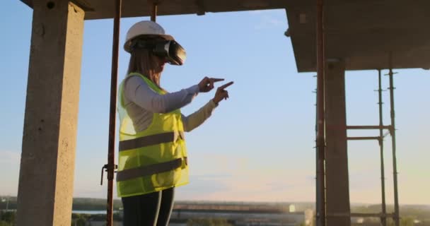 Vrメガネとヘルメットの女性検査官は、建物の計画を視覚化し、日没時に手を動かす超高層ビルの建設の進捗状況を確認します. - 映像、動画