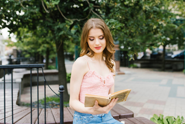 Romantic image of a young girl walking on a summer street in the Park with a bouquet of roses, city recreation and walk. Sitting on a bench with a book in his hands, reading - Photo, Image