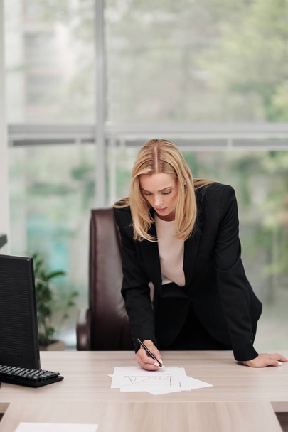 Young attractive Caucasian blond woman in black business suit sits at desk in bright office. Studying paper documents, looking at camera and smiling. - Фото, зображення