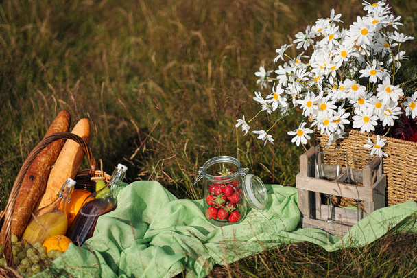 Summer picnic in the meadow on the green grass. Fruit basket, juice and bottled wine, watermelon and bread baguettes. White tablecloth and a bouquet of field daisies. - Photo, Image