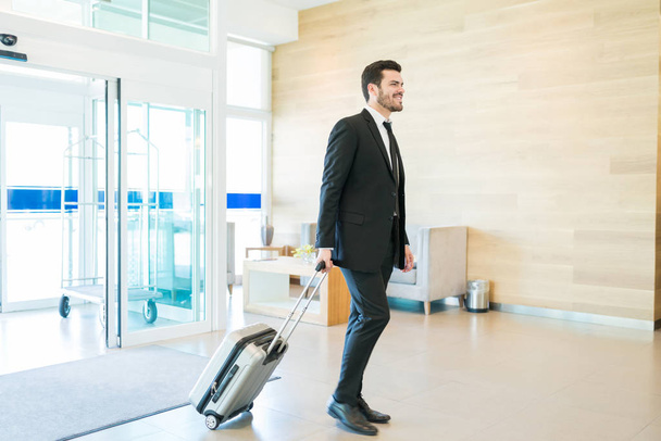 Full length of smiling Hispanic male entrepreneur pulling luggage while arriving at hotel lobby - Φωτογραφία, εικόνα