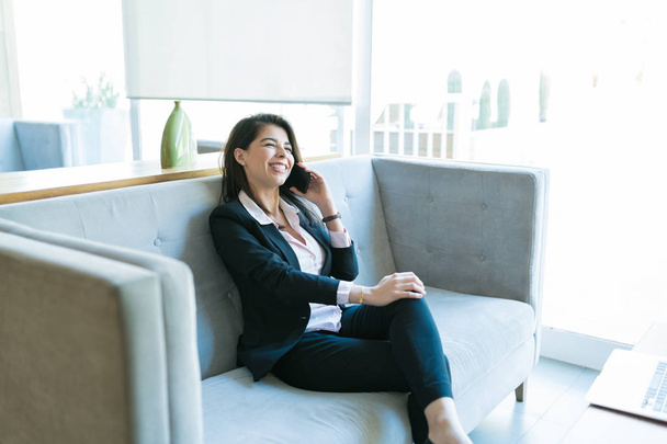 Беззаботная латиноамериканка-профессионалка сидит на диване, разговаривая по мобильному телефону с клиентом в холле
 - Фото, изображение