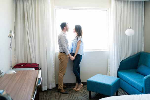 Visão lateral de comprimento total do casal recém-casado adulto médio de mãos dadas e falando enquanto estava à janela no quarto
 - Foto, Imagem