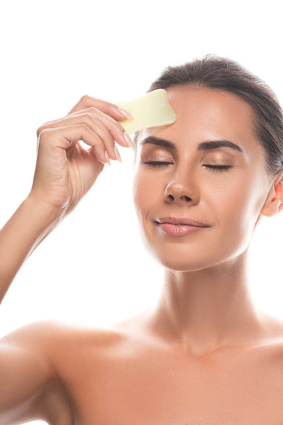 nude young woman using gua sha scraper with closed eyes isolated on white - Photo, Image