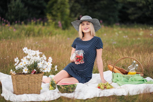 Young attractive woman in a blue dress at an outdoor picnic. A basket with daisies, watermelon, strawberries and a glass of lemonade. - Foto, imagen