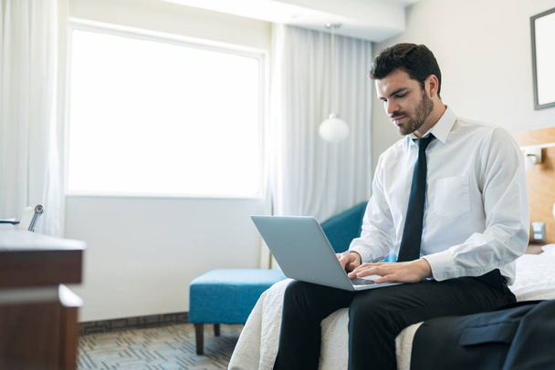 Πολυάσχολος ενήλικος επιχειρηματίας να πάρει κάποια δουλειά που γίνεται με το laptop, ενώ κάθεται στο κρεβάτι του ξενοδοχείου κατά τη διάρκεια της επαγγελματικής ταξίδι - Φωτογραφία, εικόνα