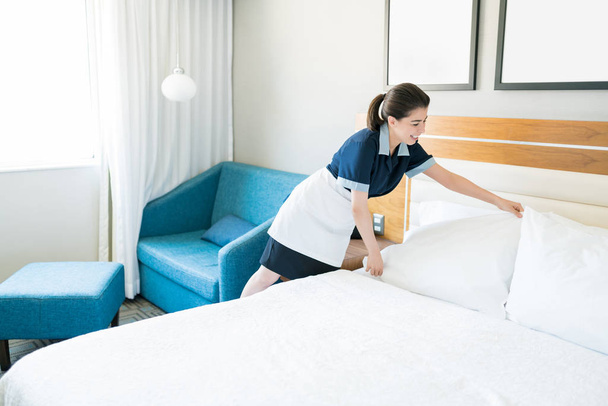 Lachende meid in uniform regelen kussens op bed in hotelkamer - Foto, afbeelding