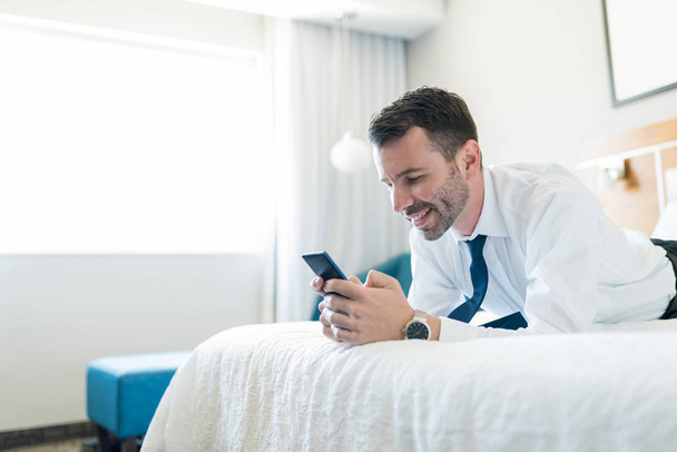 Hombre de negocios hispano adulto medio que se relaja en la cama mientras usa el teléfono celular en la habitación de hotel durante su viaje de negocios
 - Foto, Imagen