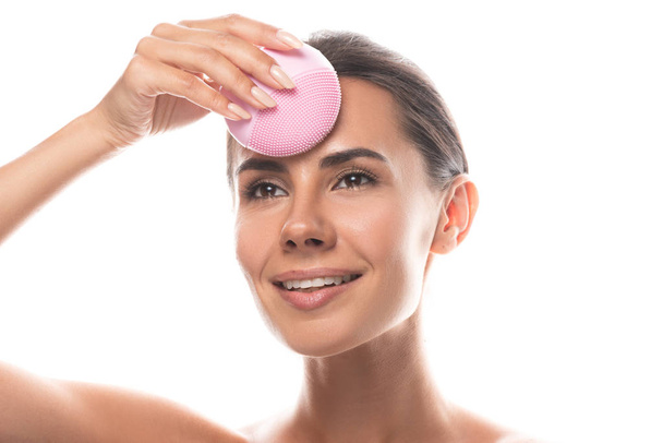 smiling young woman using facial cleansing brush and looking away isolated on white - Foto, afbeelding