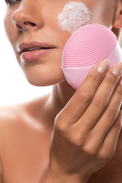 bijgesneden beeld van naakte jonge vrouw met crème op gezicht met behulp van gezichtsreiniging borstel geïsoleerd op wit - Foto, afbeelding