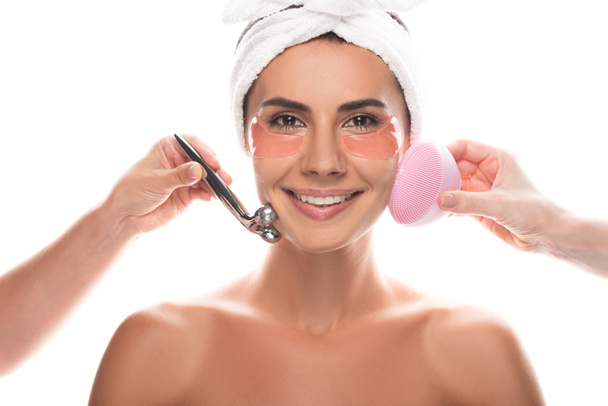 cropped view of cosmetologists using facial cleansing brush and facial massager and smiling woman with eye patches isolated on white - Photo, Image