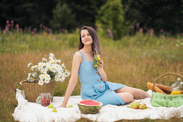 A young beautiful woman in dress is drinking lemonade from glass jar, sitting on plaid on green grass. Picnic basket, bouquet of daisies, watermelon. - Foto, imagen